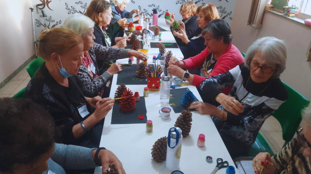 Grupo de idosos participa de atividade criativa, pintando pinhas coloridas durante uma oficina de arteterapia.