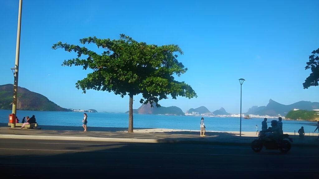 Calçadão da Praia de Icaraí em Niterói, com mar calmo, sombra e vista da orla urbana